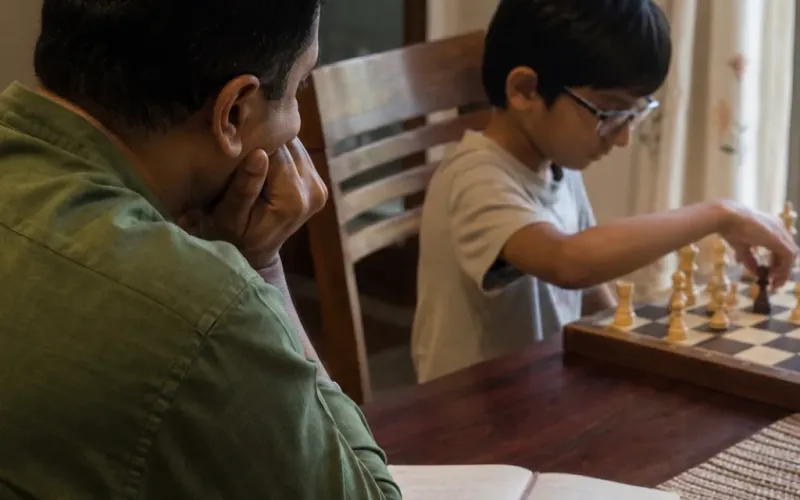 Indian parent with a face-down phone and open notebook, child at a wooden chessboard on the same table — ChessWize